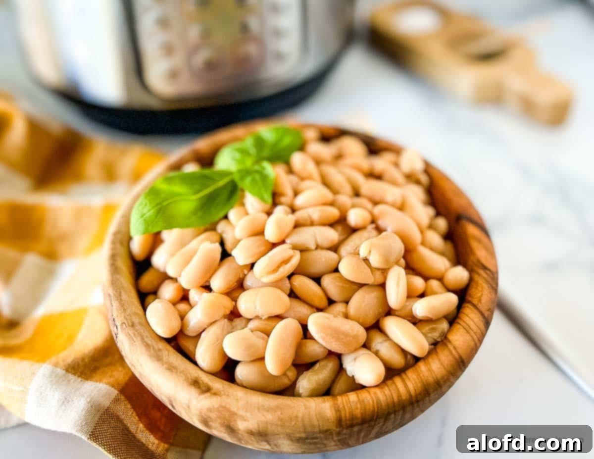 Perfectly Tender Instant Pot Cannellini Beans 9 A wooden bowl filled with pressure-cooked cannellini beans, accented with a sprig of fresh basil. A gold and white napkin and an Instant Pot are artfully blurred in the background, creating a cozy kitchen ambiance.
