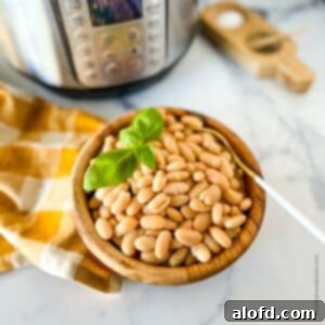 Perfectly Tender Instant Pot Cannellini Beans 10 A wooden bowl of cooked white beans, garnished with a sprig of basil. A gold and white napkin and an Instant Pot are in the background, completing the rustic kitchen scene.