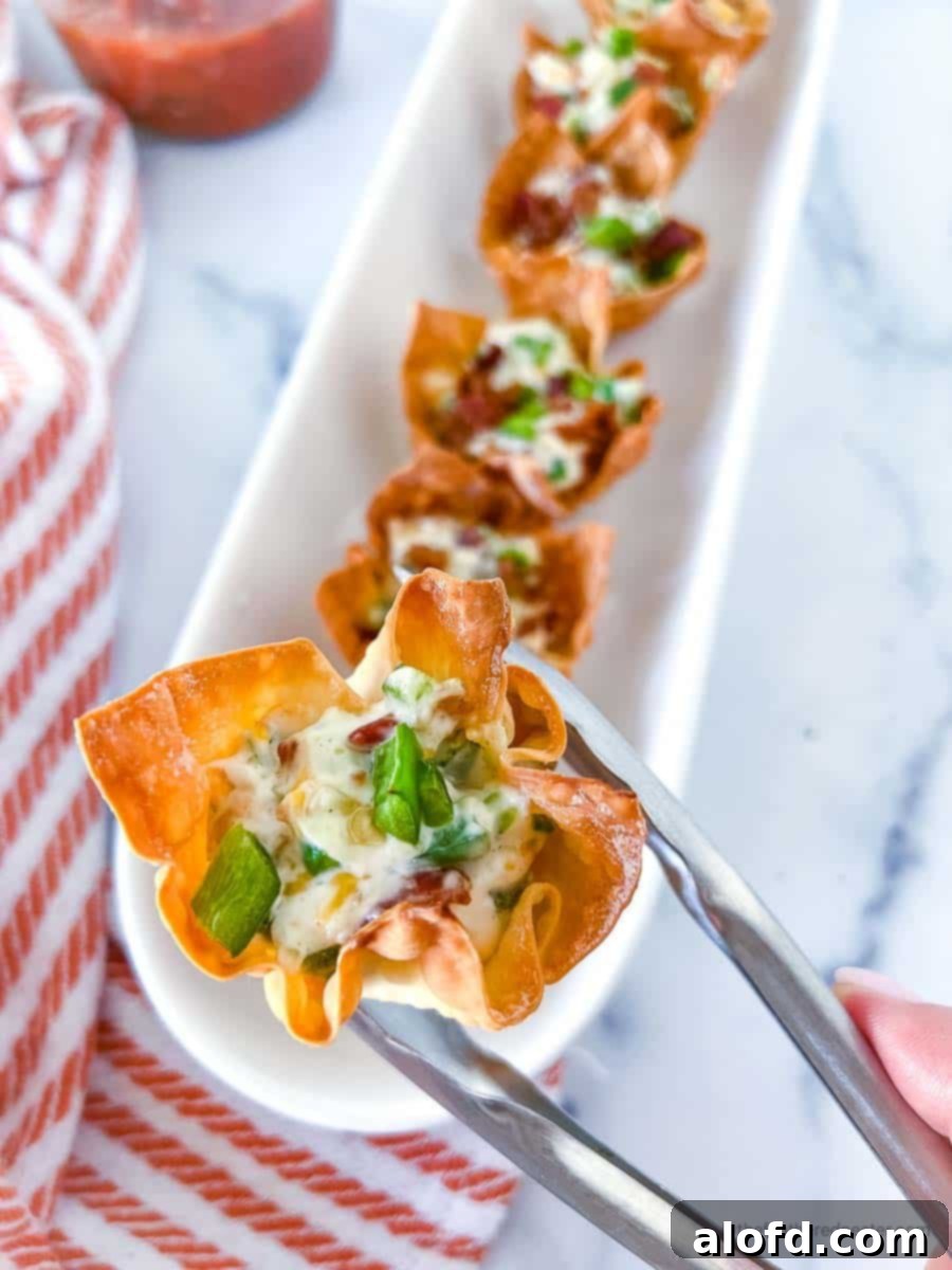 A hand and tong holding a crispy baked jalapeño popper wonton cup, with a serving tray full of similar wonton cups and a striped red cloth in the background.