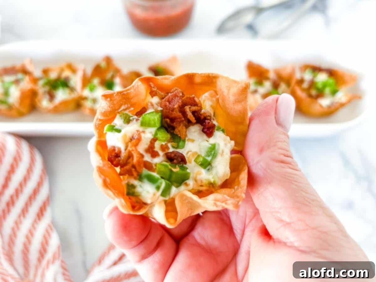 A hand holding a Jalapeno Popper Wonton Cup, with a full serving tray and a striped red cloth in the background.