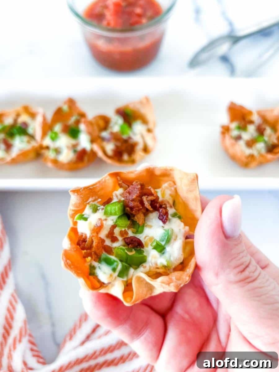 A hand holding a Jalapeno Popper Wonton Cup, with a serving tray full of crispy wonton cups, tongs, and a small bowl of dipping sauce in the background.