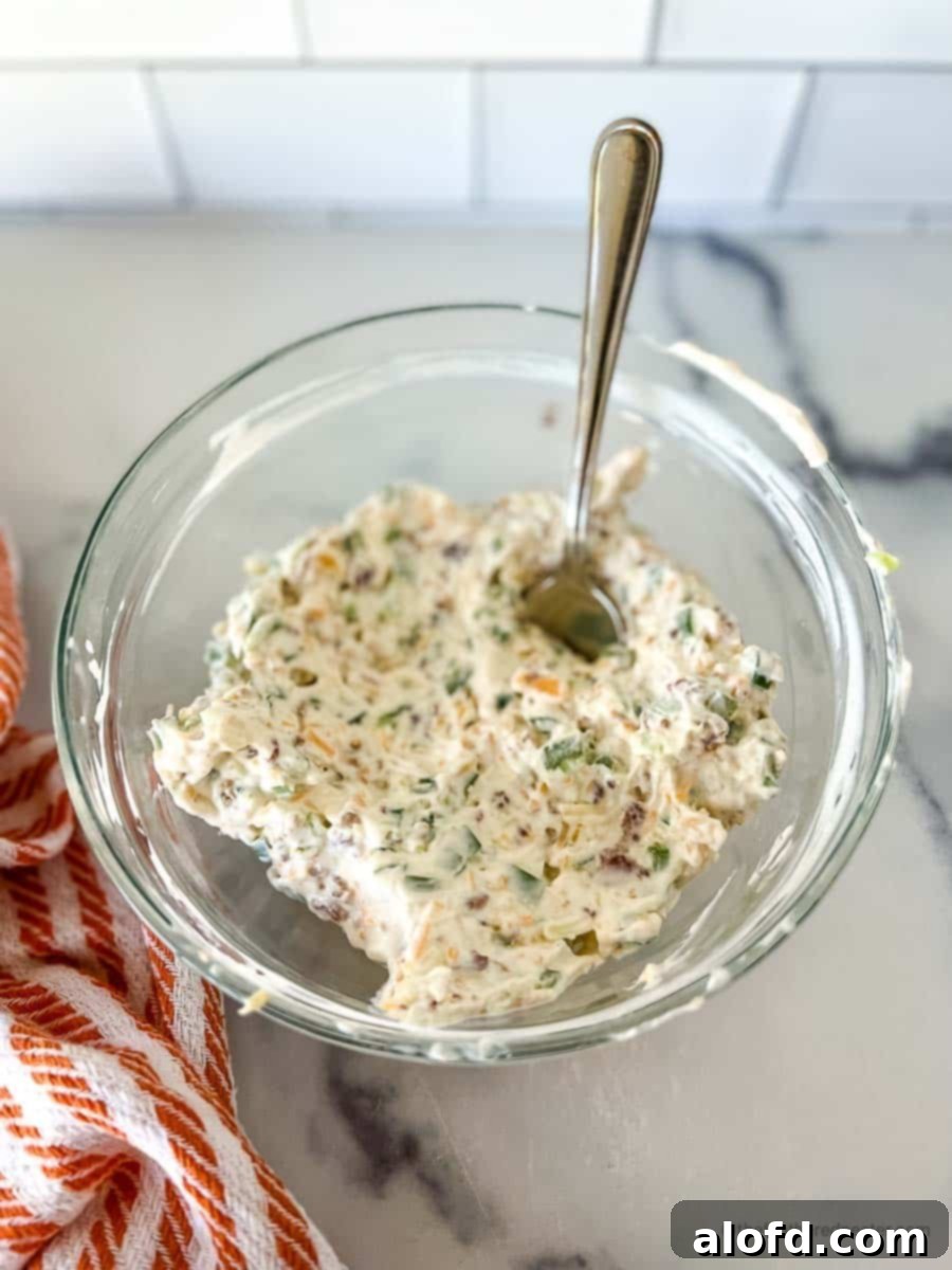 A clear bowl of jalapeño popper cheese filling with a spoon, and an orange and white napkin on the side.
