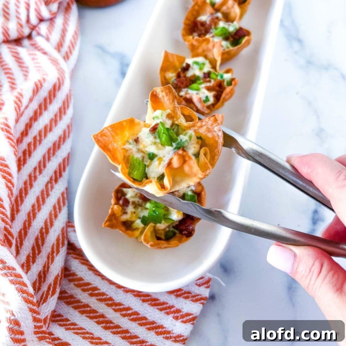A hand holding a Jalapeno Popper Wonton Cup, with a full serving tray and a striped red cloth in the background.