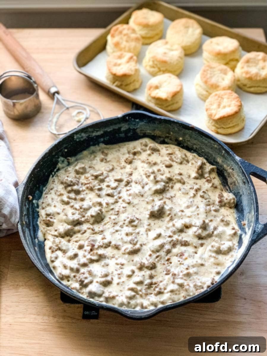 A cast iron skillet overflowing with creamy sausage gravy, with a pan of golden-brown biscuits in the soft-focus background, evoking a comforting homemade breakfast scene.