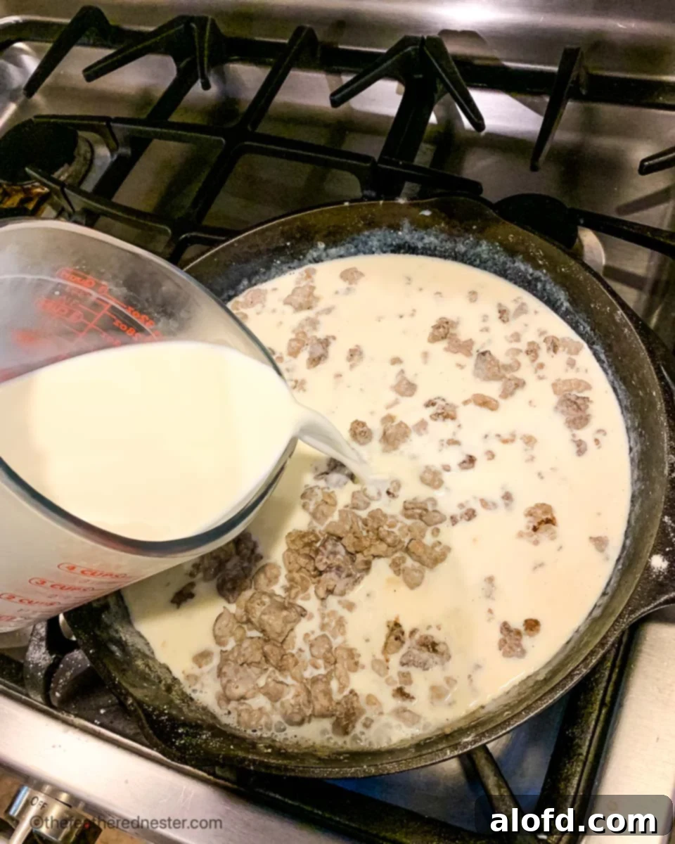 A stream of milk being poured into a cast iron skillet filled with browned sausage and flour, beginning the process of creating creamy homemade gravy.