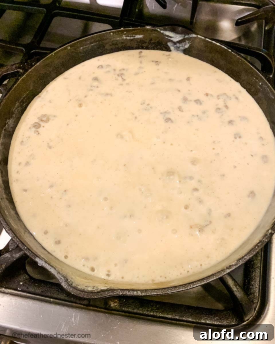 A cast iron skillet of rich, creamy sausage and milk gravy simmering on a stove burner, indicating the final stages of cooking for a hearty breakfast.
