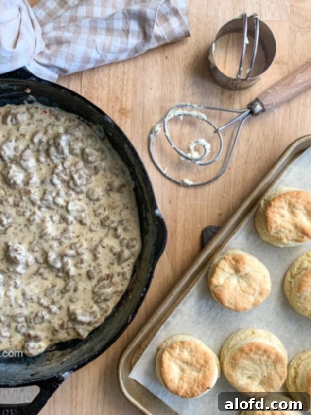 A cast iron skillet filled with savory sausage gravy, surrounded by a baking sheet of warm, golden-brown biscuits and kitchen utensils, highlighting a ready-to-serve breakfast.