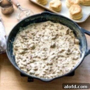 A rustic cast iron skillet filled with hearty sausage gravy, with a warm pan of golden biscuits visible in the background, signaling a cozy breakfast ready to be enjoyed.