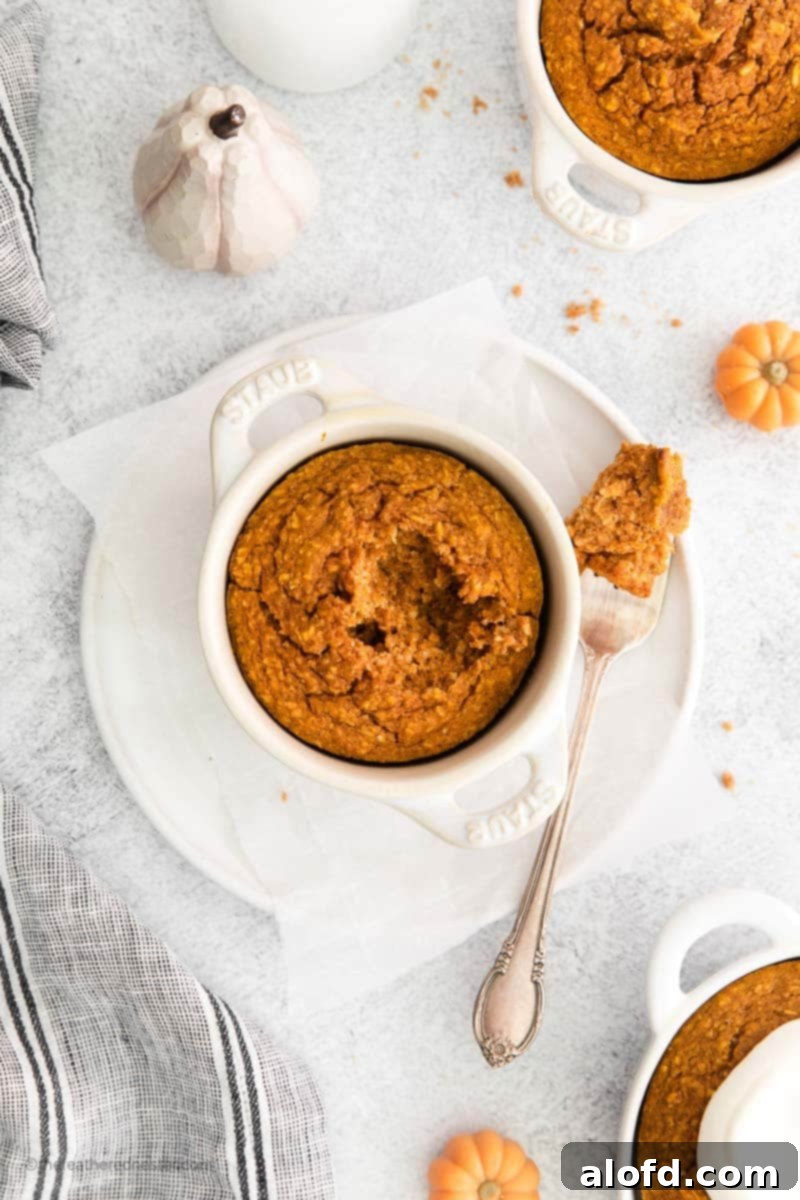 A white ramekin brimming with golden pumpkin baked oats and a fork delicately placed to the side, all arranged on a white plate. In the background, other ramekins of baked oats are softly blurred, creating a serene and inviting breakfast scene.