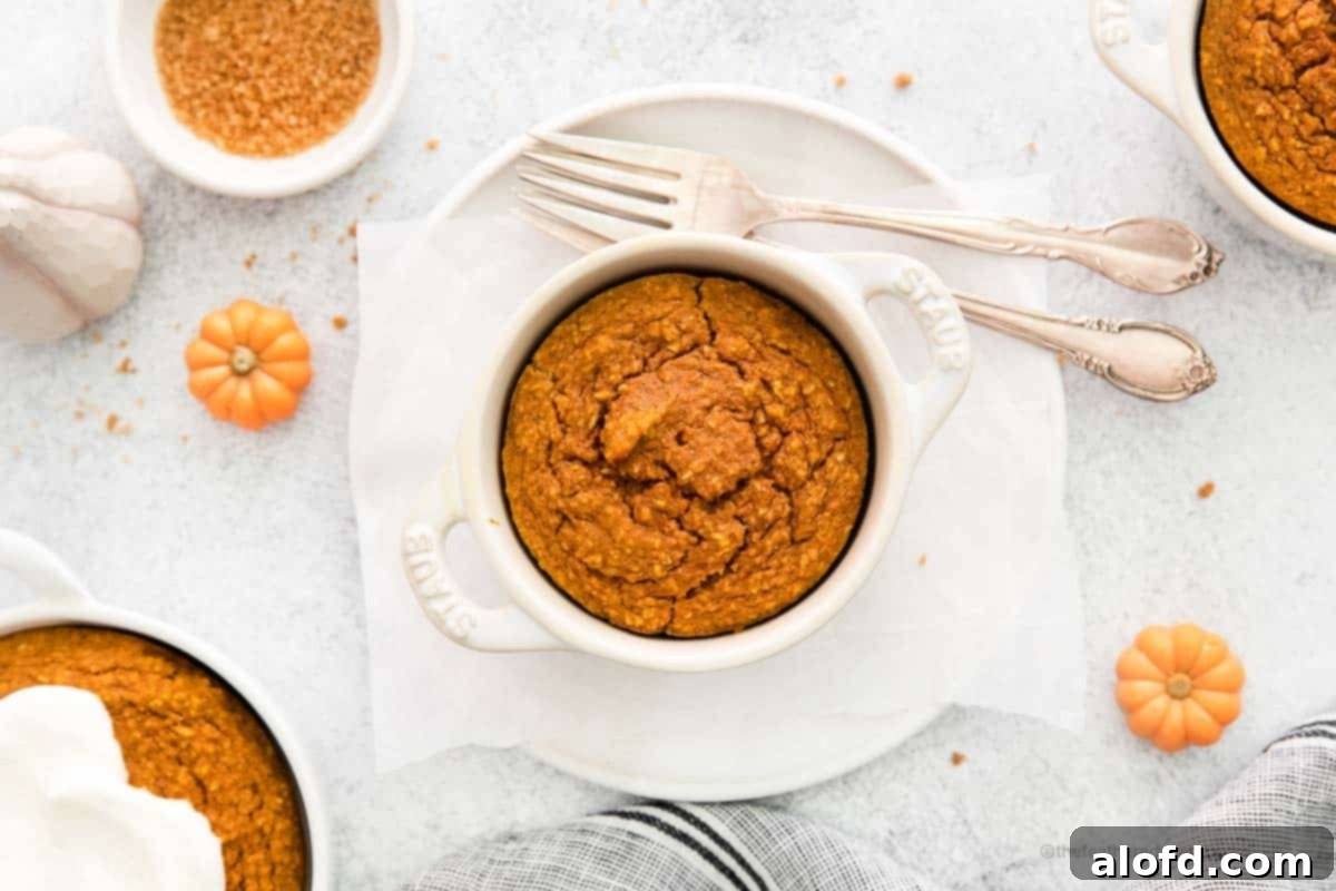 A landscape-oriented photo showcasing a single white ramekin of pumpkin baked oats, accompanied by forks, all resting on a white plate draped with a soft gray cloth. Another ramekin of oats is visible in the gently blurred background, completing a cozy breakfast scene.