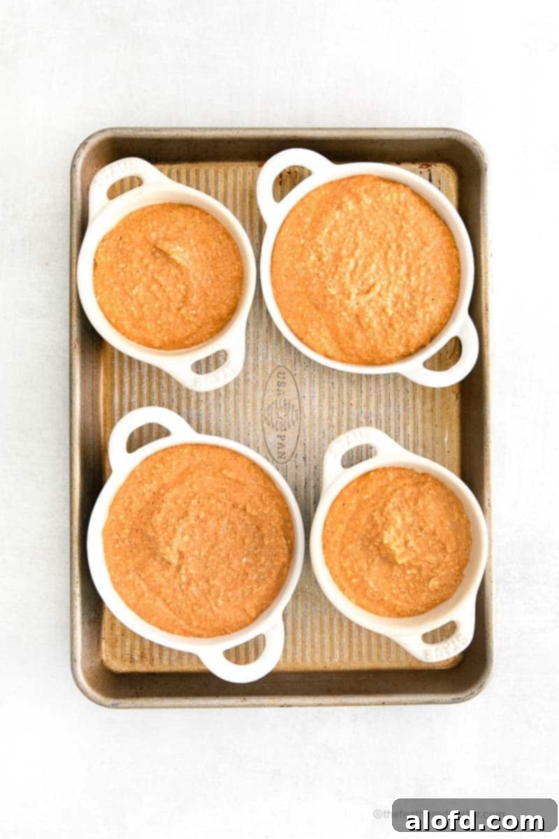 Four white ramekins are neatly arranged on a baking sheet, each filled with unbaked pumpkin spice oatmeal batter, ready to go into the oven.