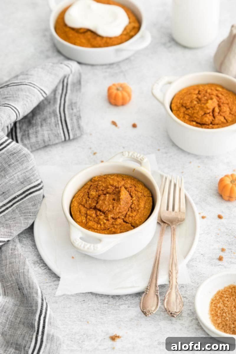 A freshly baked white ramekin of pumpkin oatmeal, perfectly golden, is presented on a white plate with two forks resting beside it. Another ramekin of baked oatmeal is blurred in the background, creating a cozy and inviting scene.