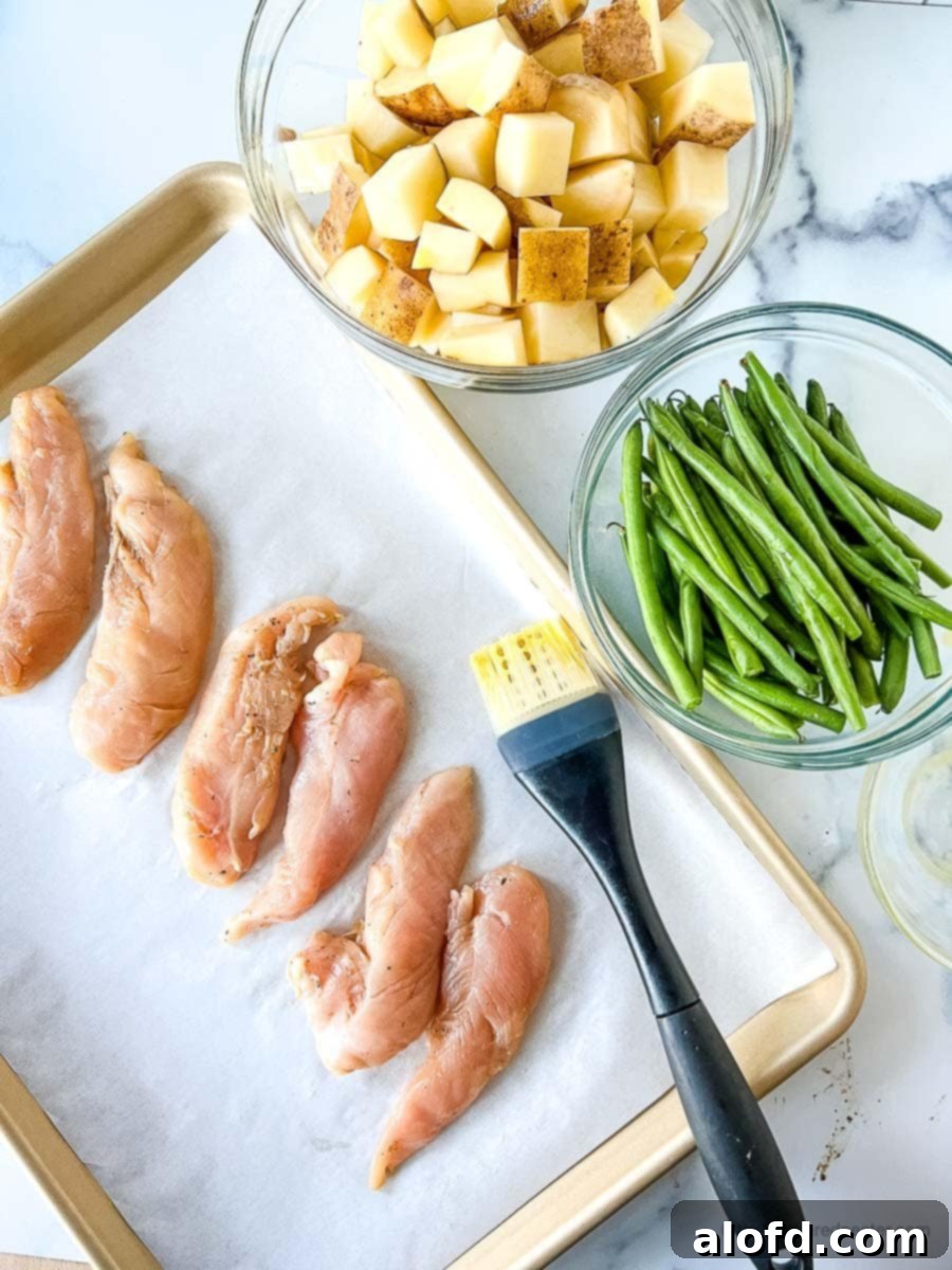 Marinated chicken tenders neatly arranged on parchment paper on a baking sheet, accompanied by a bowl of diced potatoes and asparagus, with a basting brush ready for seasoning, showcasing the prep before baking.