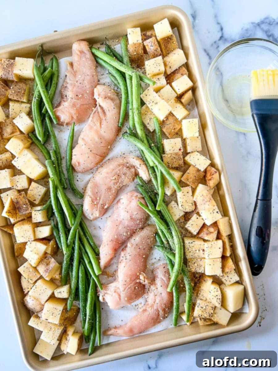 A baking sheet prepped with raw chicken tenders, asparagus, and potatoes, all lightly seasoned and drizzled with olive oil, ready for the oven, with a basting brush and oil container nearby.