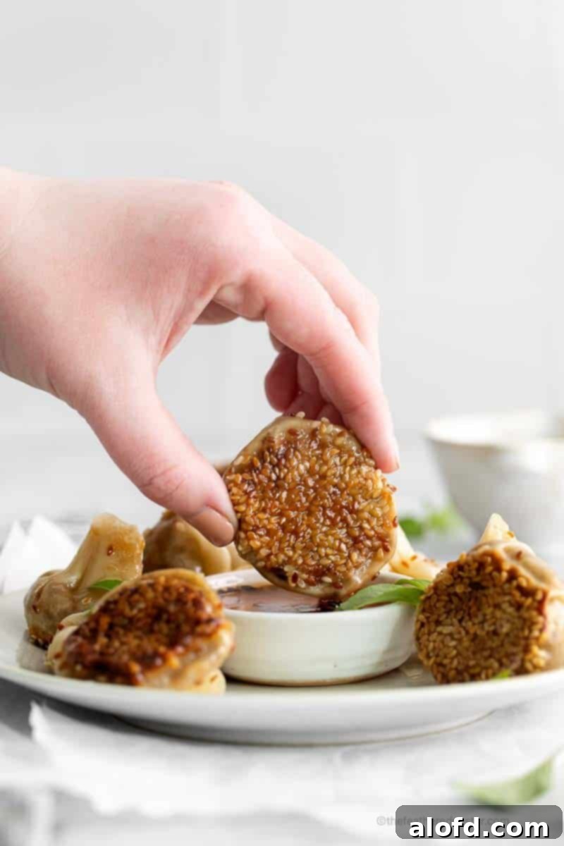 Umami Mushroom Pockets 14 A hand gently dips an Asian appetizer, generously coated in toasted sesame seeds, into a small bowl of fiery garlic chili oil, capturing a moment of delicious anticipation.