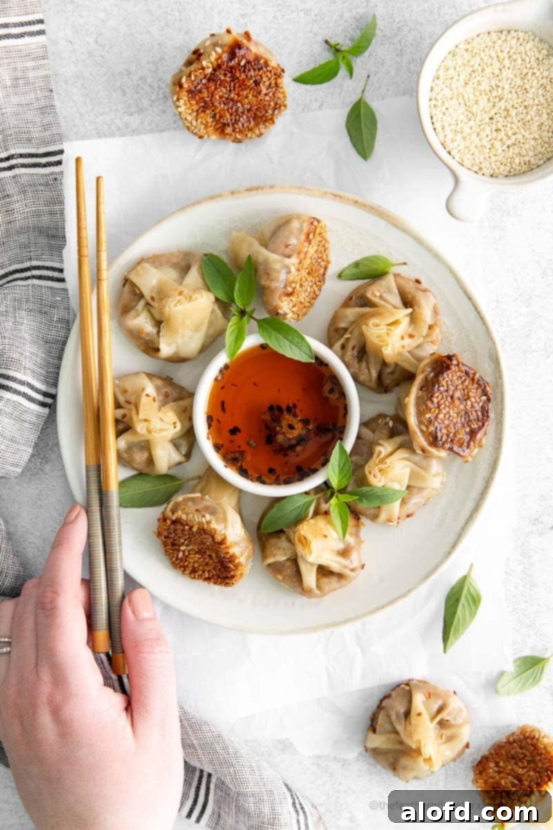 Umami Mushroom Pockets 5 A hand gracefully holds a white plate, showcasing a beautifully arranged selection of mushroom dumplings. A pair of chopsticks rests beside them, and a small bowl of fiery garlic chili oil occupies the center, inviting a delicious dip.