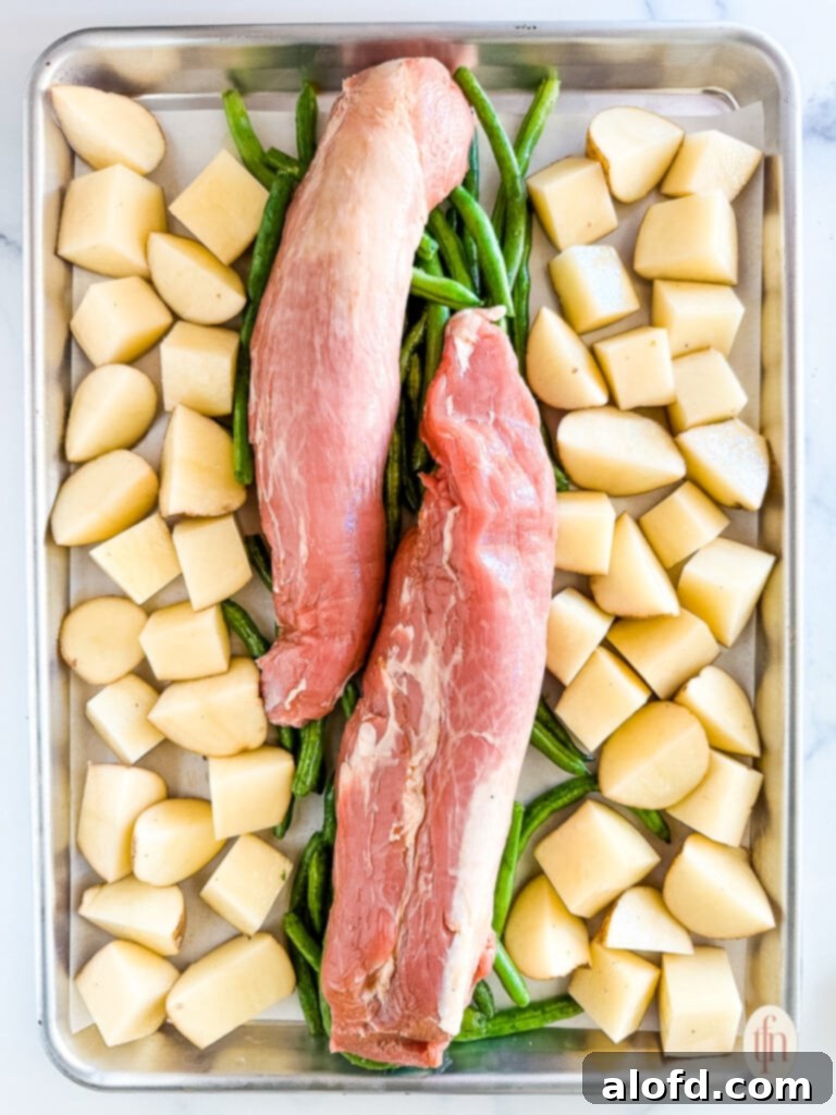 Effortless Sheet Pan Pork Tenderloin 6 Two pieces of uncooked meat on a baking pan with diced white potatoes and string beans.