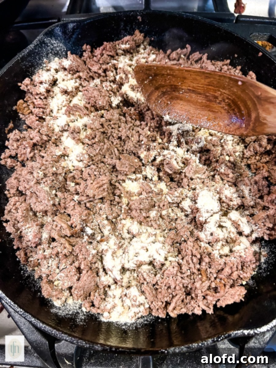 Browning ground beef with onion and garlic powder in a cast iron skillet, a crucial step for the Cheeseburger Tater Tot Casserole.