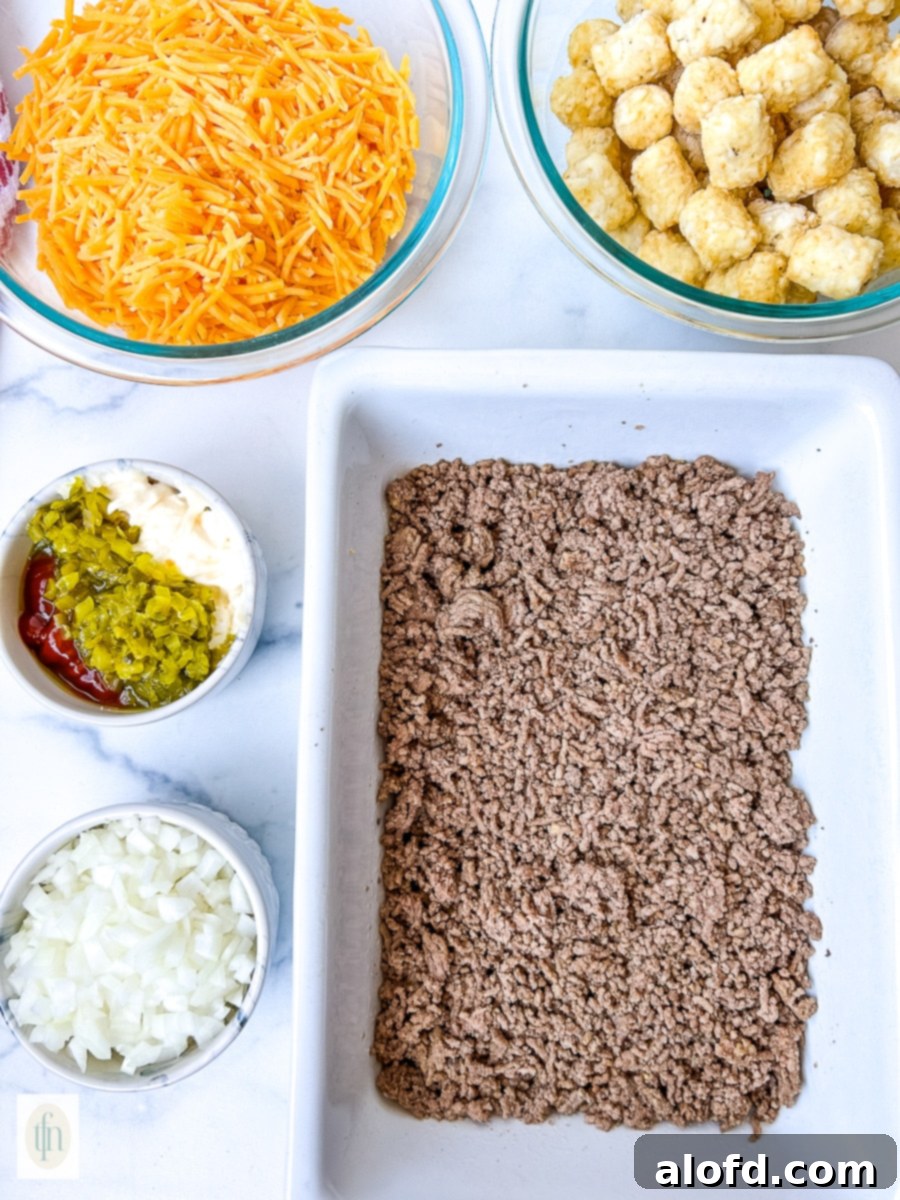 Cooked ground beef in a white baking dish surrounded by smaller bowls of diced onion, bell pepper, shredded cheese, and tater tots, ready for assembly.