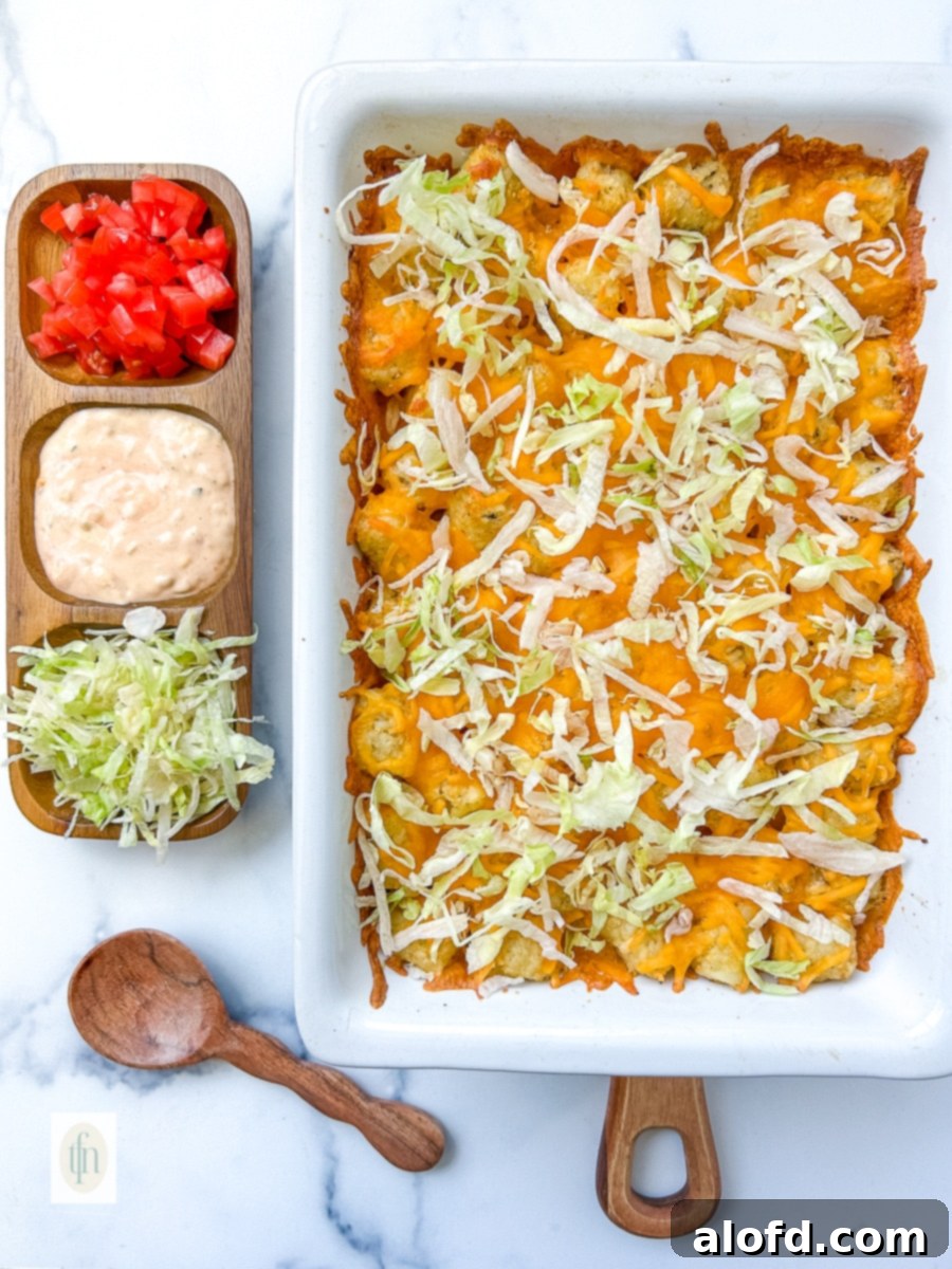 A hamburger potato casserole topped with shredded lettuce, alongside a bowl of diced tomatoes and a generous serving of homemade Big Mac sauce, ready for garnishing.