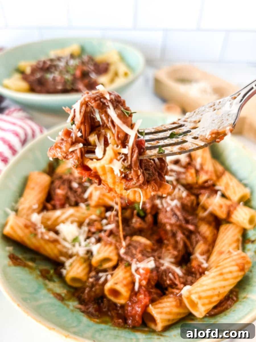 Braised Short Rib Rigatoni 3 A silver fork lifting a piece of rigatoni coated in rich braised short rib ragu. In the background, two green bowls of short rib rigatoni, a red striped cloth, and salt and pepper shakers create an inviting scene.