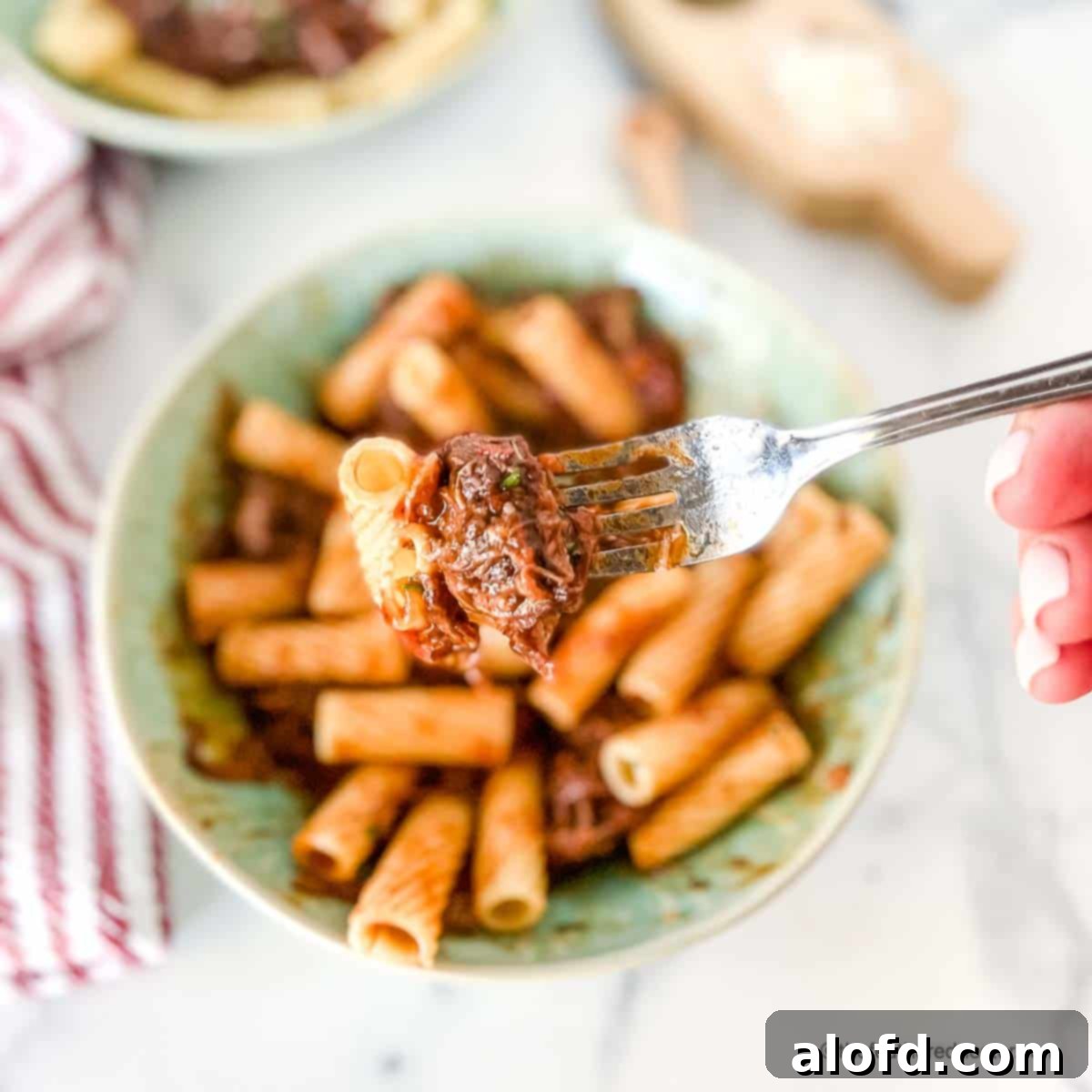 Braised Short Rib Rigatoni 4 A fork holding pasta generously coated with braised short rib ragu, with a green bowl of short rib rigatoni visible in the soft focus background.