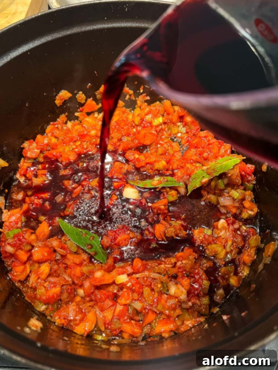 Braised Short Rib Rigatoni 8 Pouring dry red wine into the sautéed vegetables and tomato paste in a Dutch oven, initiating the deglazing process for the braised short rib ragu.