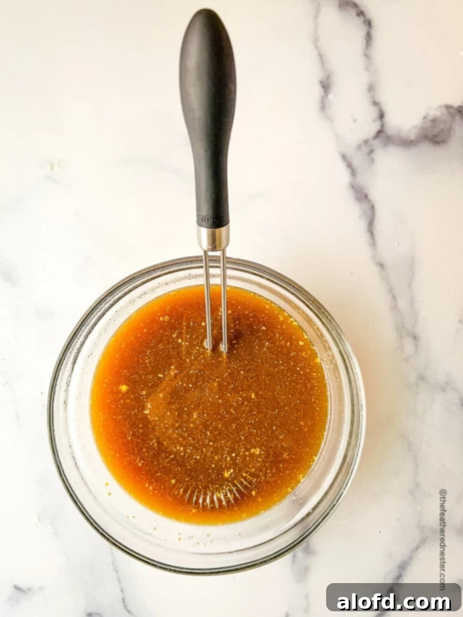 A small, clear glass bowl holds a rich, brown Dr. Pepper ham glaze, with a whisk resting inside, ready for application. The glaze appears thick and inviting.
