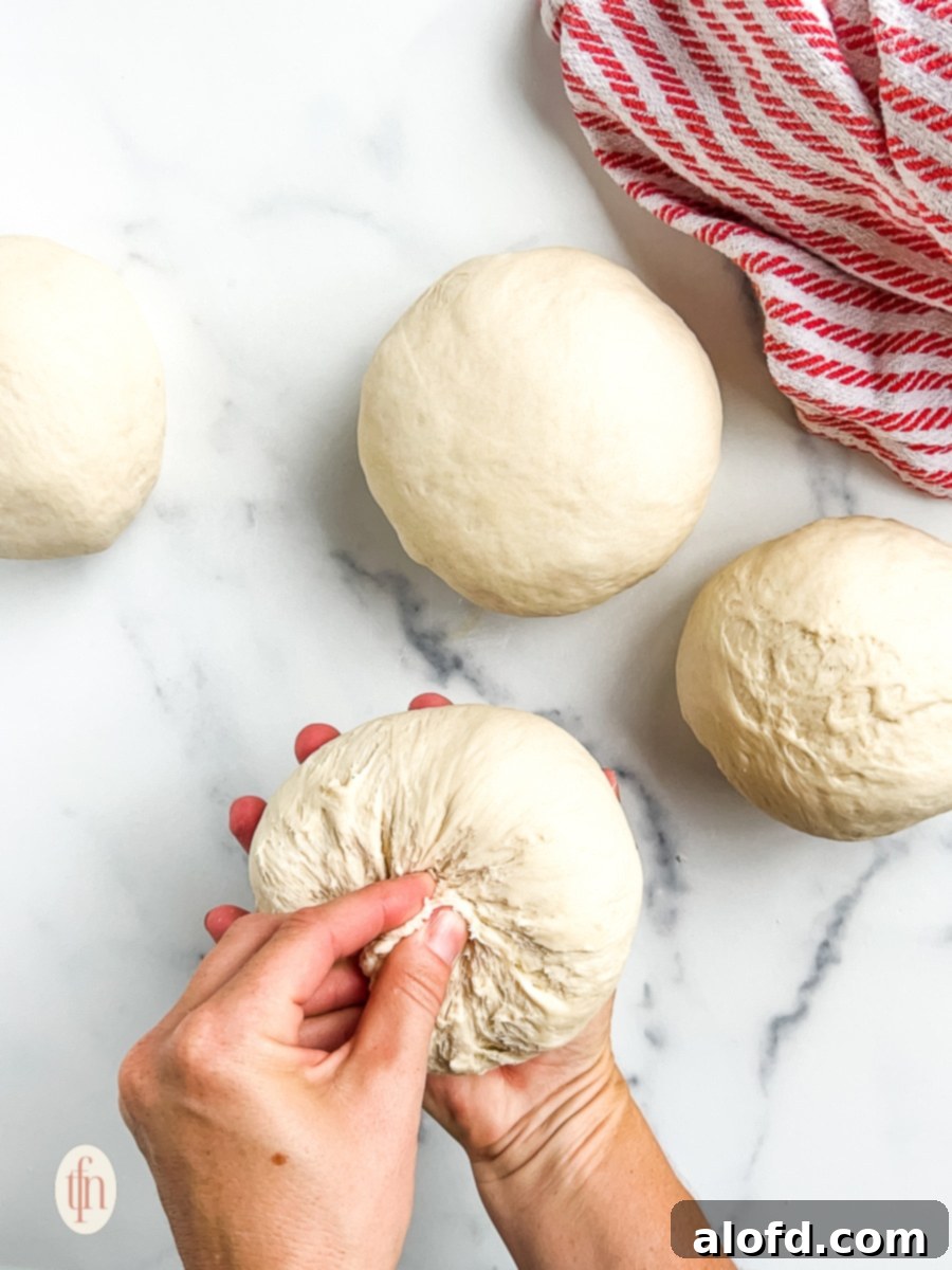 Artisan Sourdough Soup Bowls 9 A hand gently pinching and shaping the bottom of a smooth, rounded piece of sourdough dough on a lightly floured surface, preparing it for its final rise.