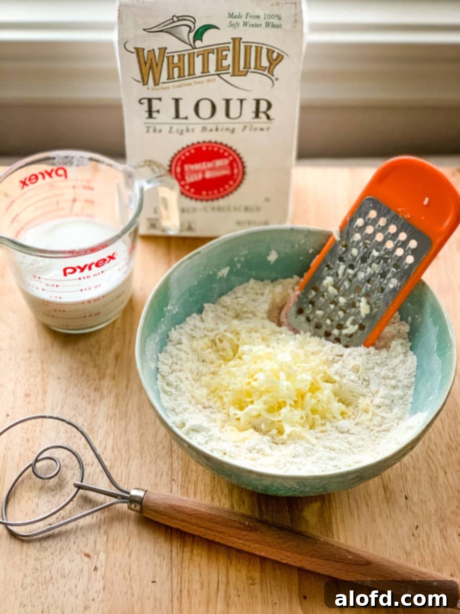 Blue bowl of grated butter and self rising flour with buttermilk, a bag of self rising flour, and a dough whisk shown.