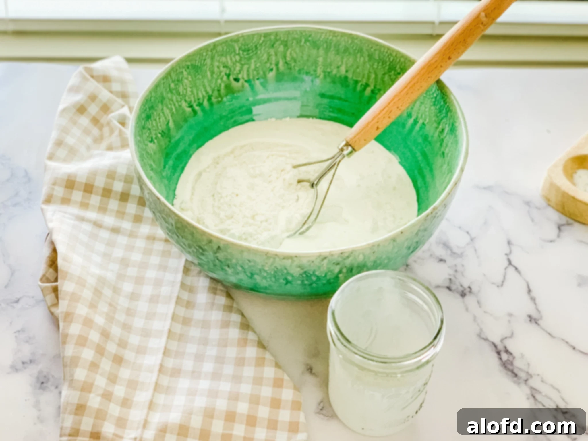 A green bowl with flour and a whisk with a jar and kitchen towel at the side.