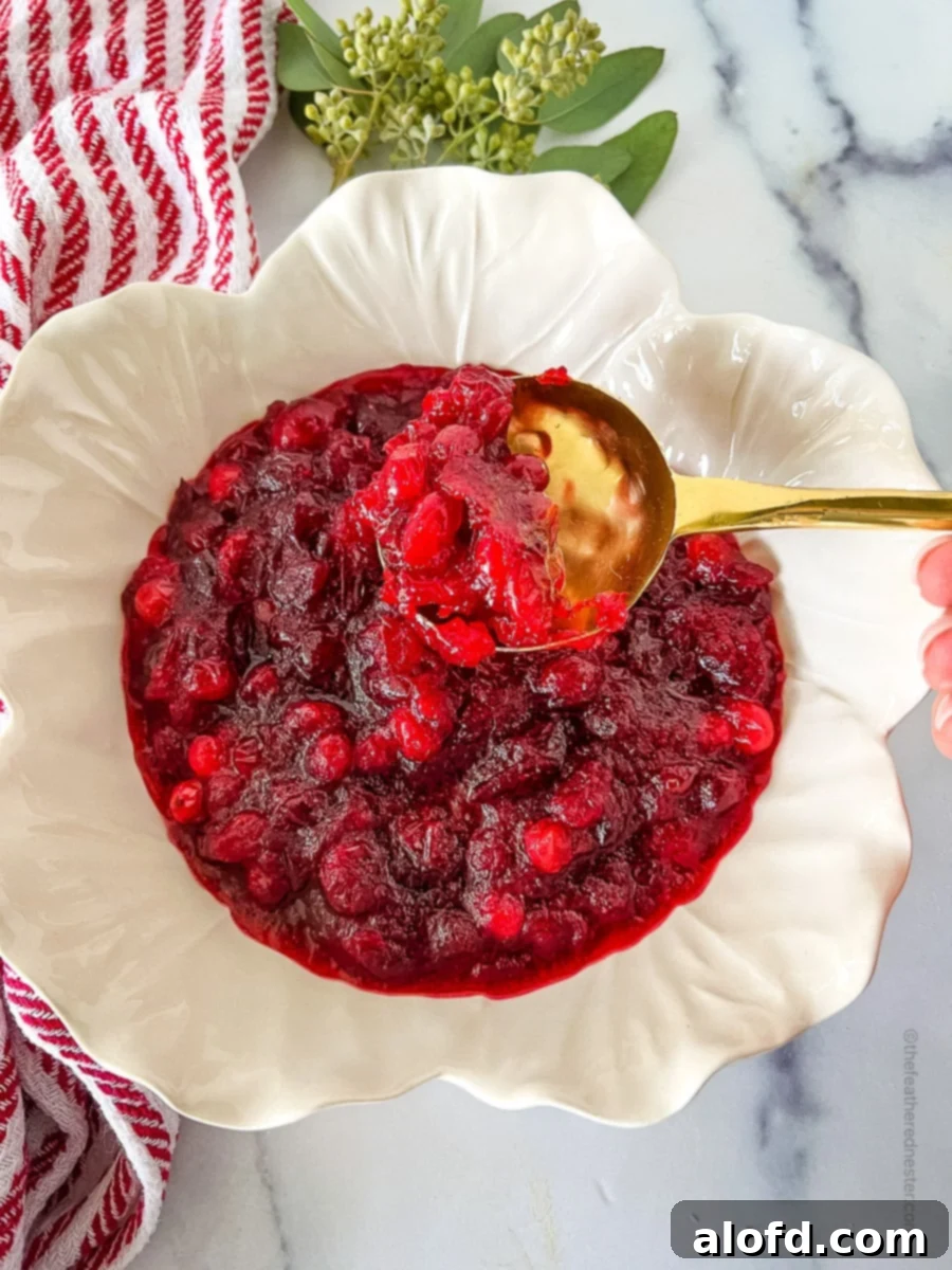 A white bowl of glistening homemade cranberry sauce with a gold serving spoon, accompanied by a festive red and white napkin, creating a cozy holiday scene.