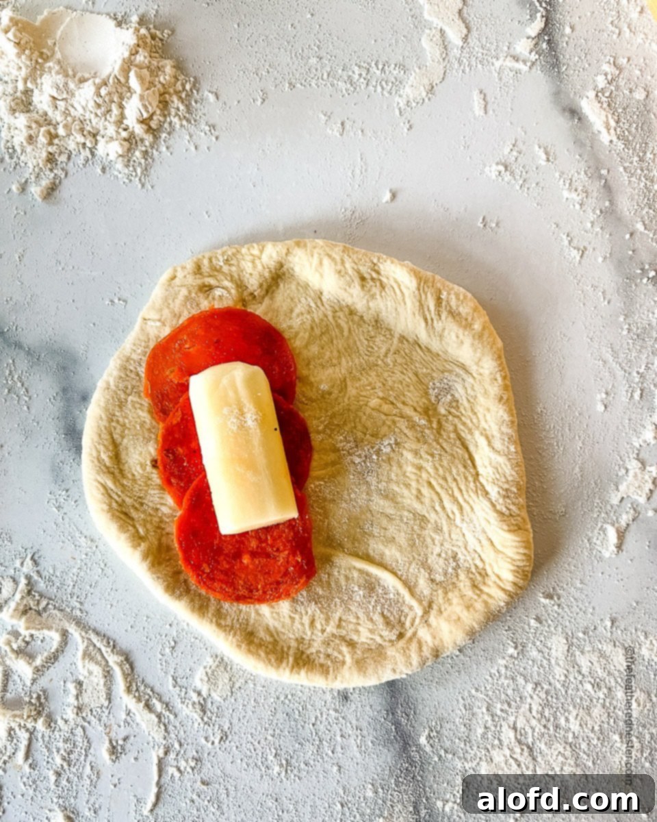 Savory Pepperoni Calzone Pockets 11 A flattened dough circle with three pepperoni slices, a piece of string cheese, and a dollop of marinara sauce on one half.