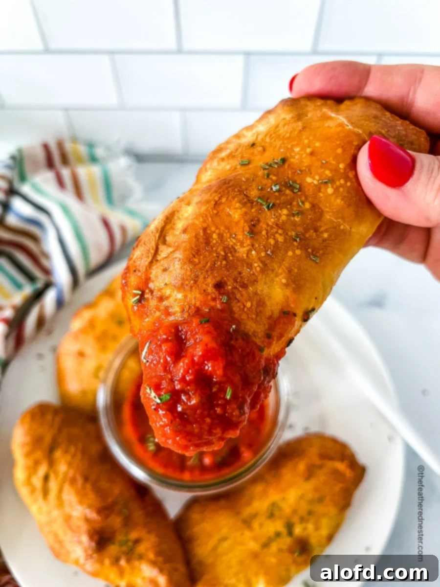 Savory Pepperoni Calzone Pockets 14 A hand holding a dipped mini pepperoni calzone, with a plate full of calzones in the background, ready to be enjoyed.