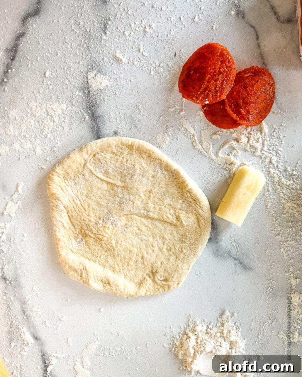 Savory Pepperoni Calzone Pockets 10 A flattened dough circle with a cut piece of string cheese and three pepperoni slices placed neatly to the side, ready for filling.