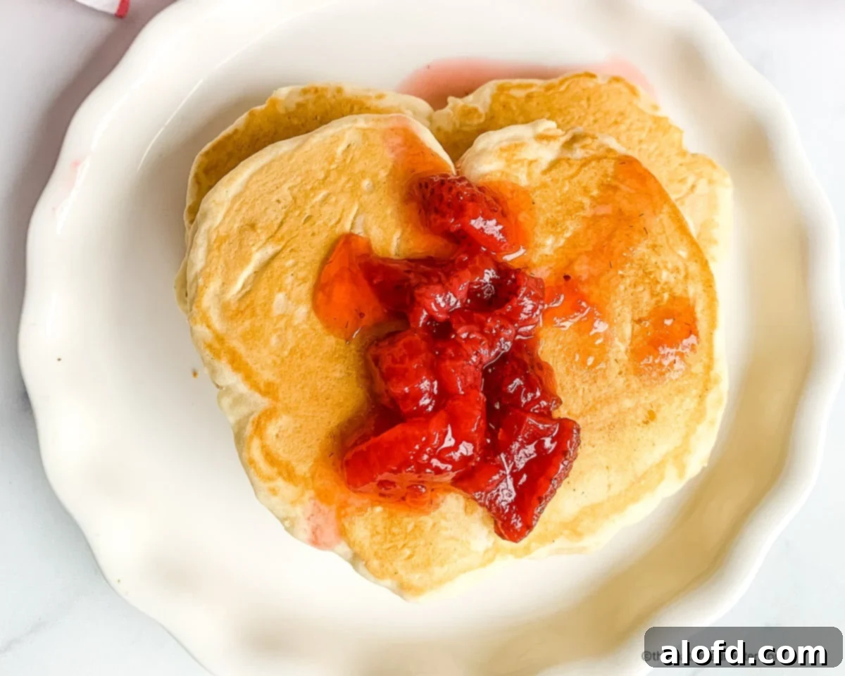 Plated serving of heart shaped pancakes with homemade strawberry syrup. Start your Valentine's Day with love by making heart-shaped pancakes for two! This delicious and cozy breakfast is perfect for your sweetheart, especially when served with strawberry pancake syrup. A sweet way to kick off your special day together!