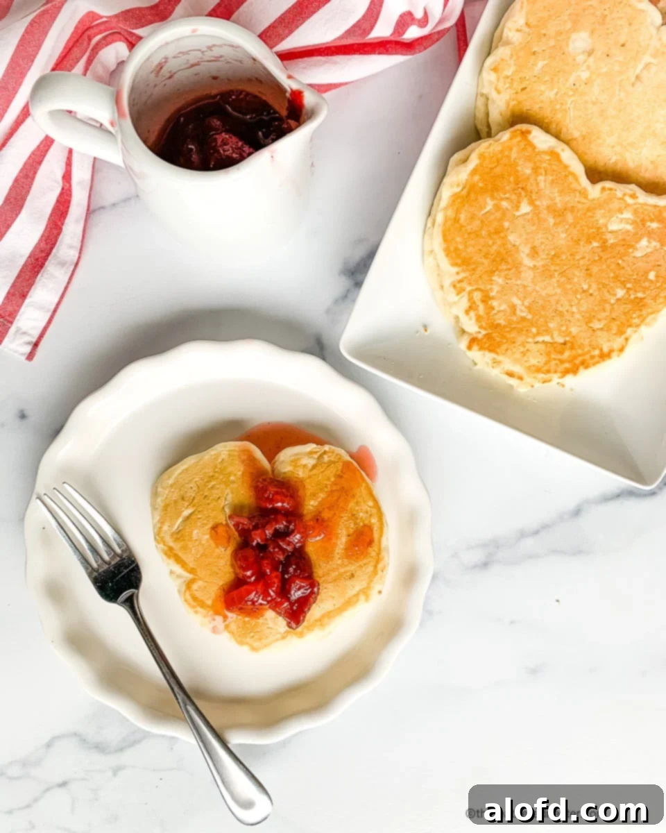 Heart shaped pancakes on a white platter next to a plated short stack topped with strawberry pancake syrup. Start your Valentine's Day with love by making heart-shaped pancakes for two! This delicious and cozy breakfast is perfect for your sweetheart, especially when served with strawberry pancake syrup. A sweet way to kick off your special day together!