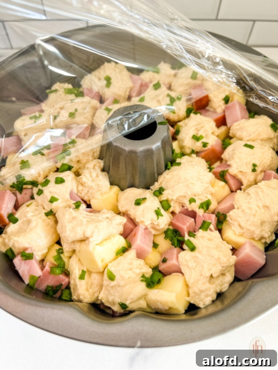 Raw monkey bread with ham and brie on a pan with plastic wrap on top.