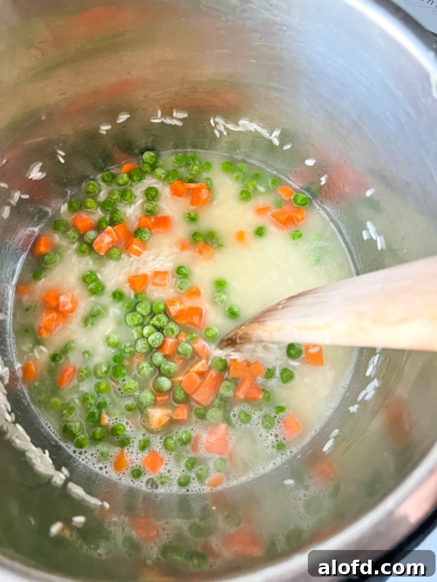 Hot broth being poured into an Instant Pot containing sautéed rice and frozen mixed vegetables, ready for pressure cooking.