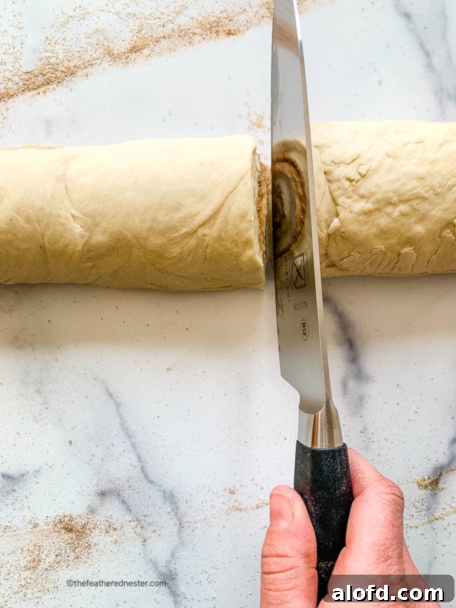Fluffy Sourdough Discard Cinnamon Rolls 13 A chef's knife carefully slicing into a log of sourdough discard cinnamon roll dough on a marble countertop, starting from the center.