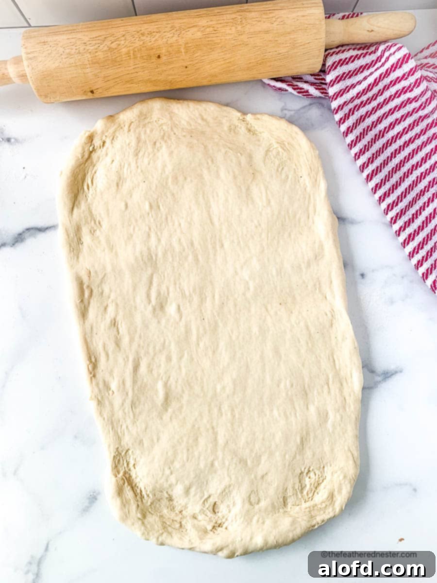 Fluffy Sourdough Discard Cinnamon Rolls 8 A large piece of sourdough discard dough on a marble countertop next to a rolling pin and a red striped kitchen towel, ready for shaping.