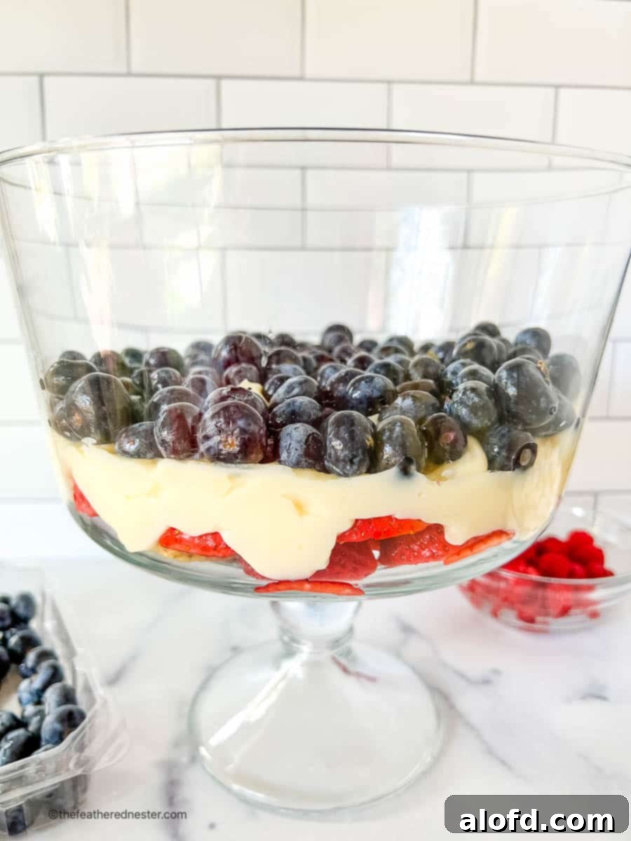Close-up shot of hands meticulously assembling a cheesecake salad, layering fresh fruit and creamy pudding in a large glass trifle bowl, showcasing the process.