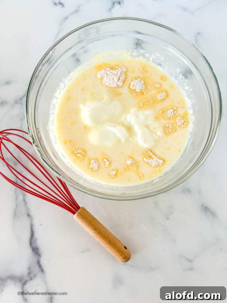 A red whisk sits beside a clear mixing bowl, which contains a blend of yogurt, dry pudding mix, and milk, illustrating the initial steps of preparing the cheesecake filling.