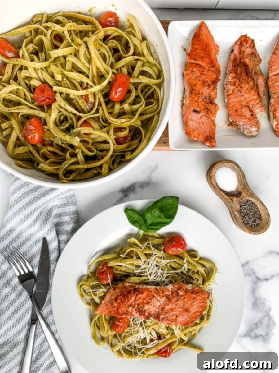 Bright Salmon Pesto Pasta 5 A close-up shot of a dinner plate featuring salmon pesto pasta, garnished with fresh basil and parmesan, with a blurred background of a larger bowl of pasta and a platter of cooked fish.