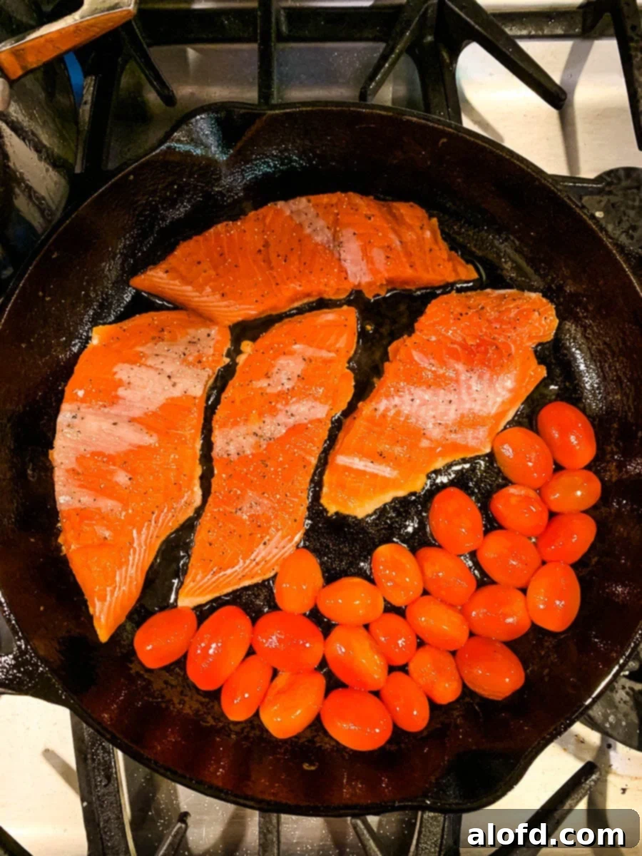 Bright Salmon Pesto Pasta 7 A cast-iron skillet shows salmon fillets searing alongside blistered cherry tomatoes, demonstrating the simultaneous cooking process.