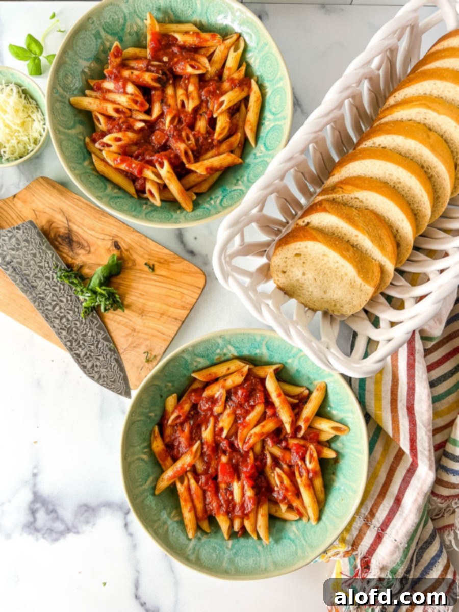 Two plated servings of penne pasta with meatless tomato basil pasta sauce next to a white basket with sliced French bread, ready for serving.