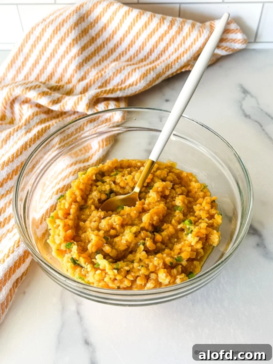 A small, clear glass dish filled with vibrant orange-red cooked lentils, accompanied by a white serving spoon, highlighting their soft texture.