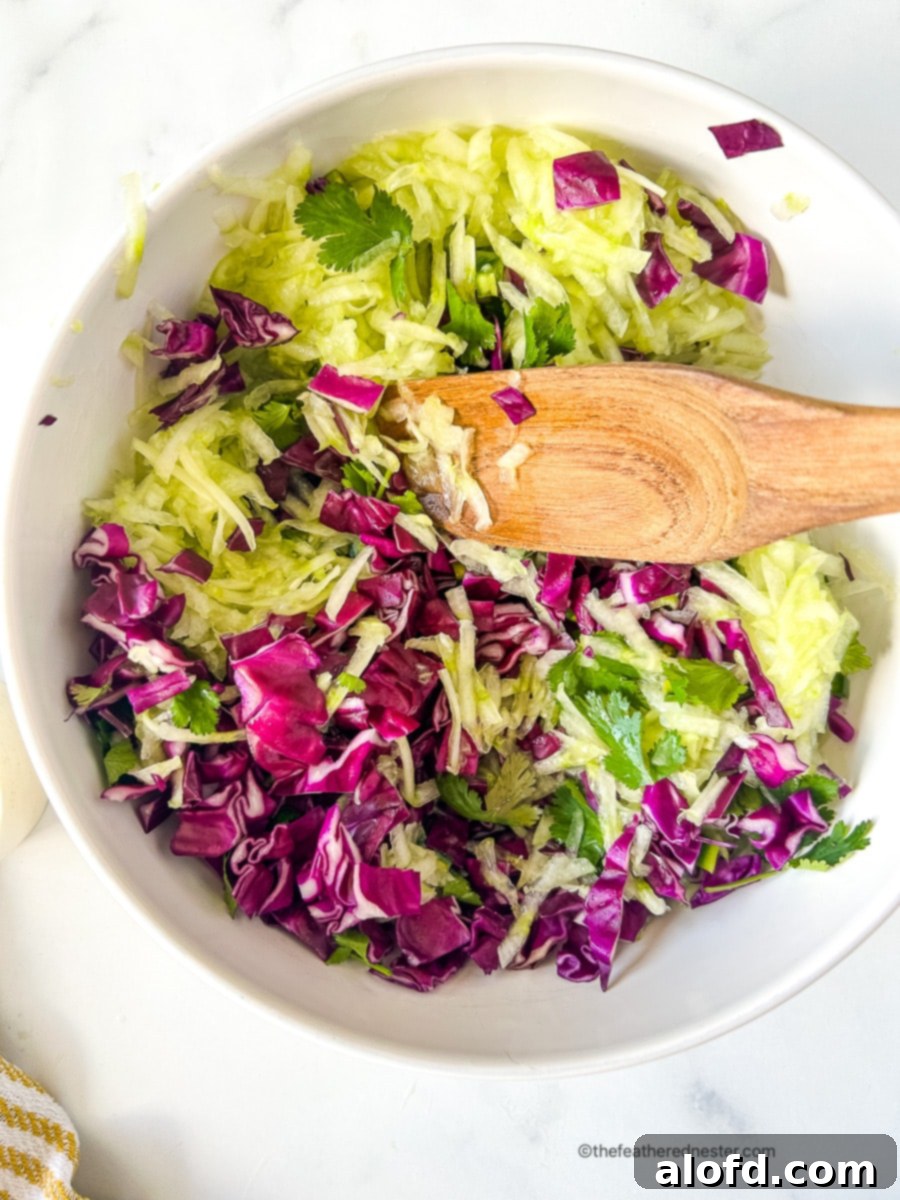 Using wooden spoon to combine shredded cucumbers and chopped red cabbage in a mixing bowl.