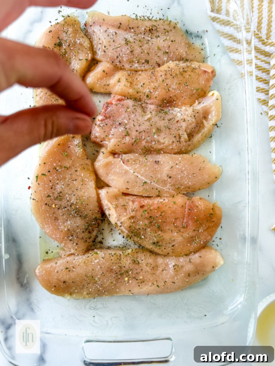 A hand sprinkling salt and pepper over raw chicken cutlets, preparing them for seasoning and baking.