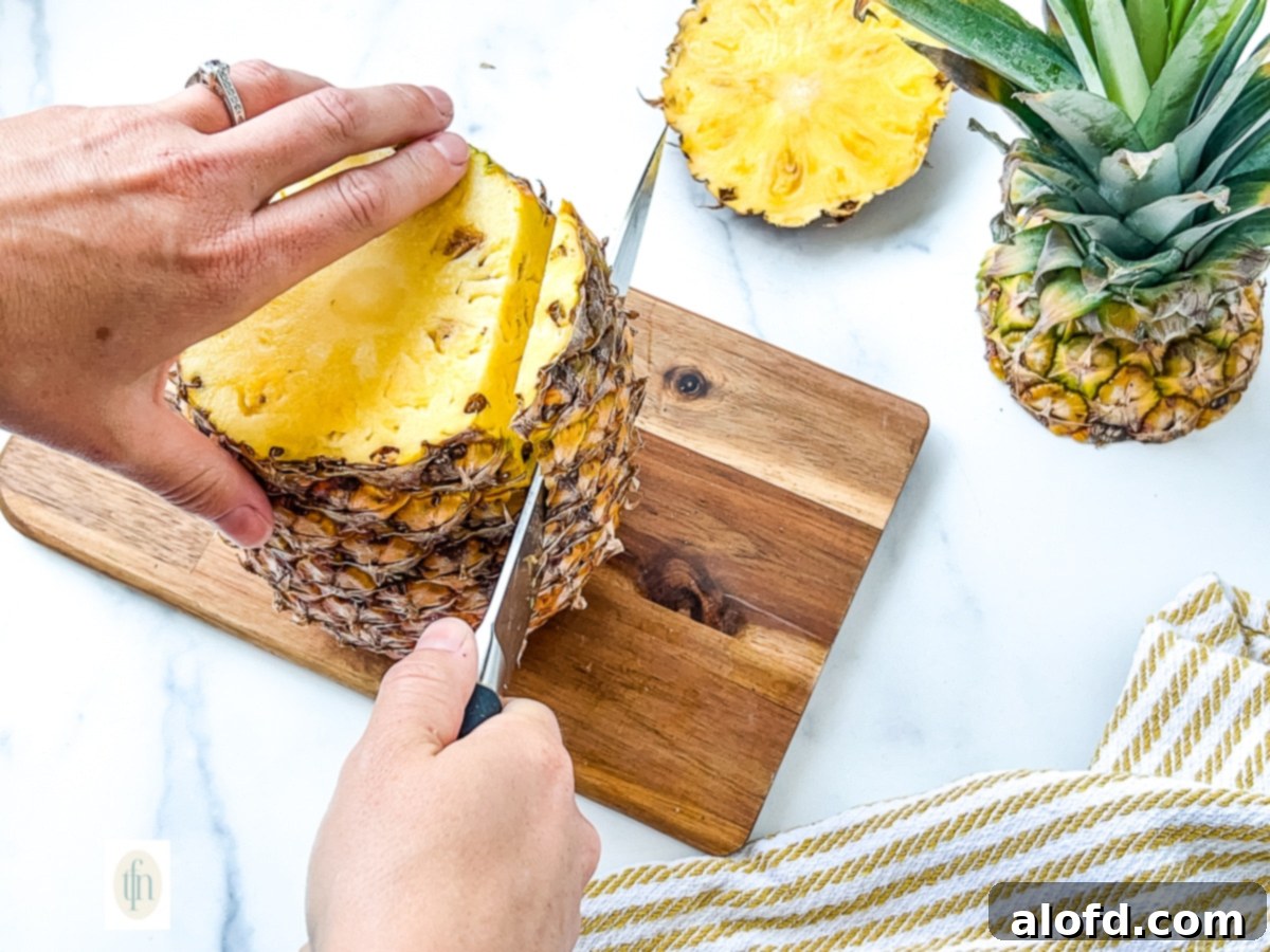 A person using a large kitchen knife to carefully slice off the prickly skin of a pineapple.